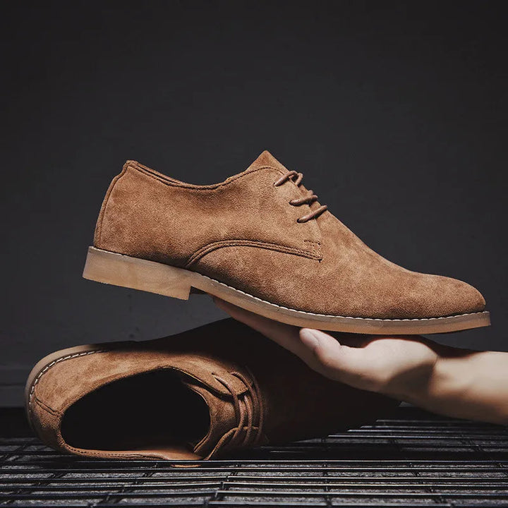 Stack of three pairs of men's suede Oxford shoes in different colors: brown, navy, and gray.