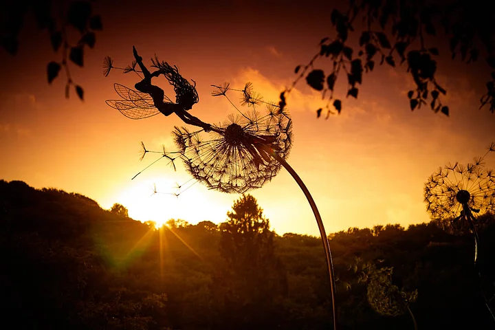 Whimsical metal fairy with lifelike dandelion stakes, ideal for enchanting garden decor, adding magic to outdoor spaces.