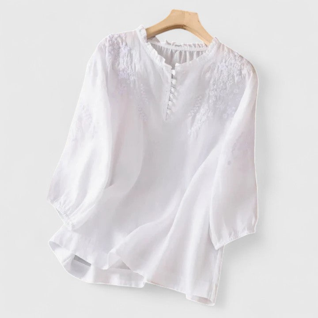 Women's white embroidered blouse with floral patterns, elegant ruffle collar, and playful pom-pom trim. Three-quarter sleeves.